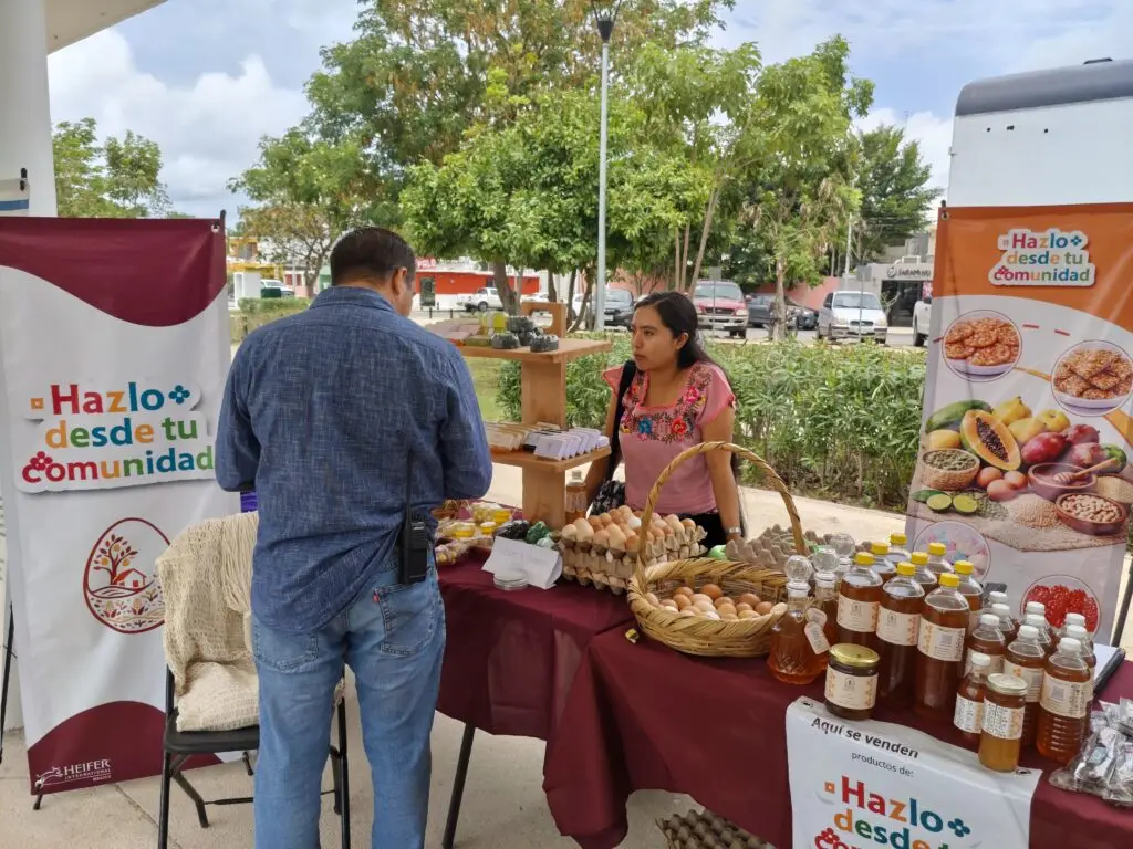 Emprendedora rural interactúa con un visitante en una feria comunitaria. Sobre la mesa cubierta con manteles guindas se exhiben alimentos frescos y productos hechos a mano bajo el letrero "Hazlo desde tu comunidad".