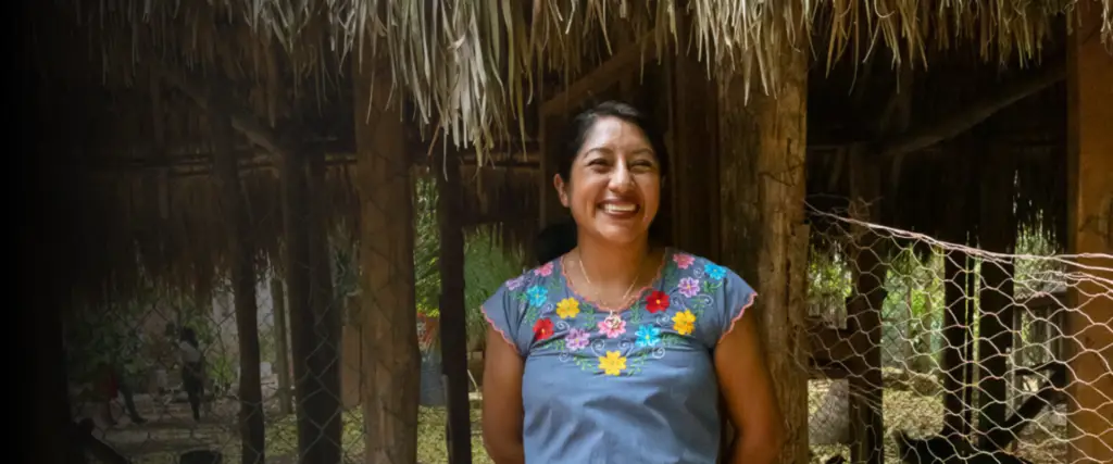 Mujer emprendedora sonriendo frente a un gallinero de traspatio donde cría gallinas ponedoras como parte de un proyecto de producción avícola comunitaria.